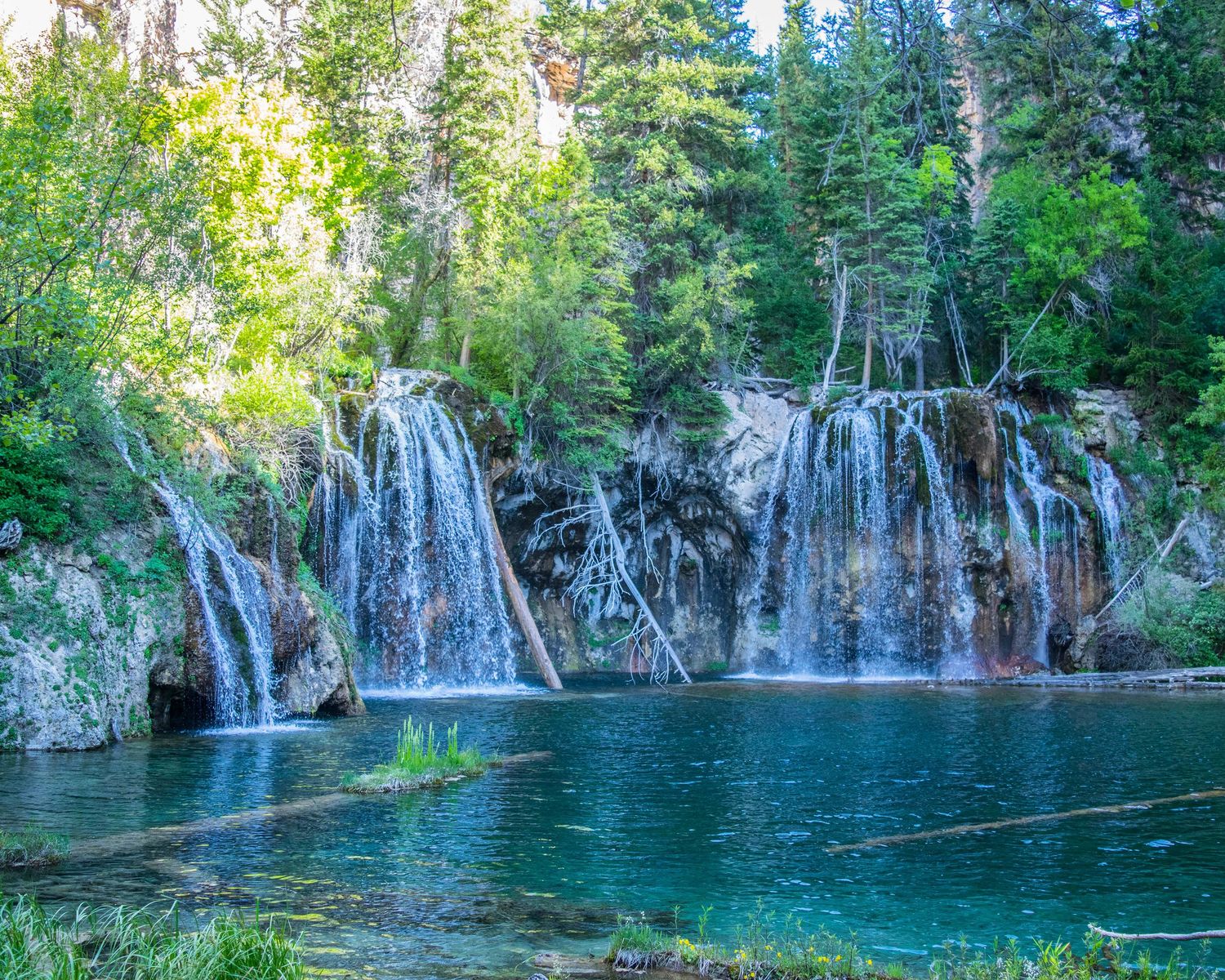 Stunning turquoise pool reflects multiple cascading waterfalls surrounded by lush green trees at Hanging Lake, Colorado.