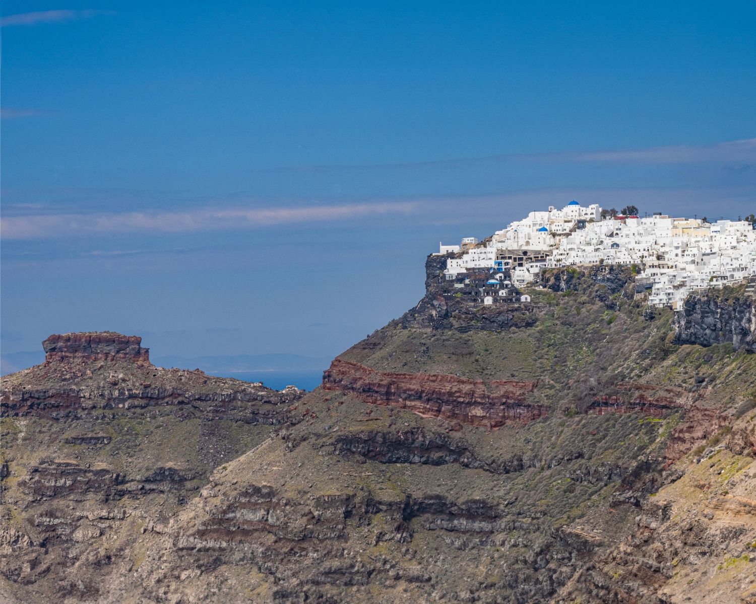 White-washed buildings perched atop dramatic red volcanic cliffs in Santorini, Greece, under a clear blue sky.