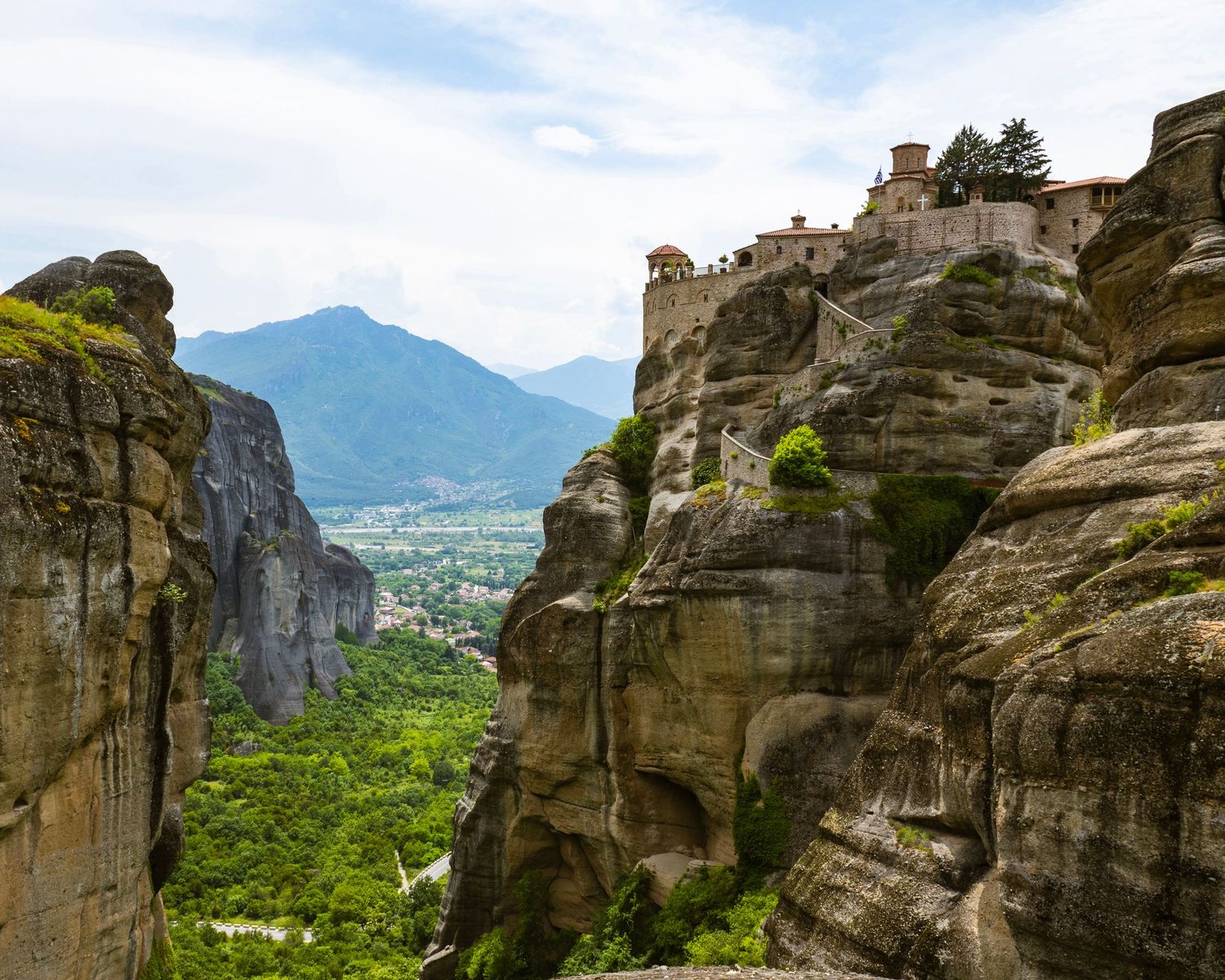 Meteora monasteries perched atop dramatic rock formations in Greece with lush green valleys and mountains in the background.