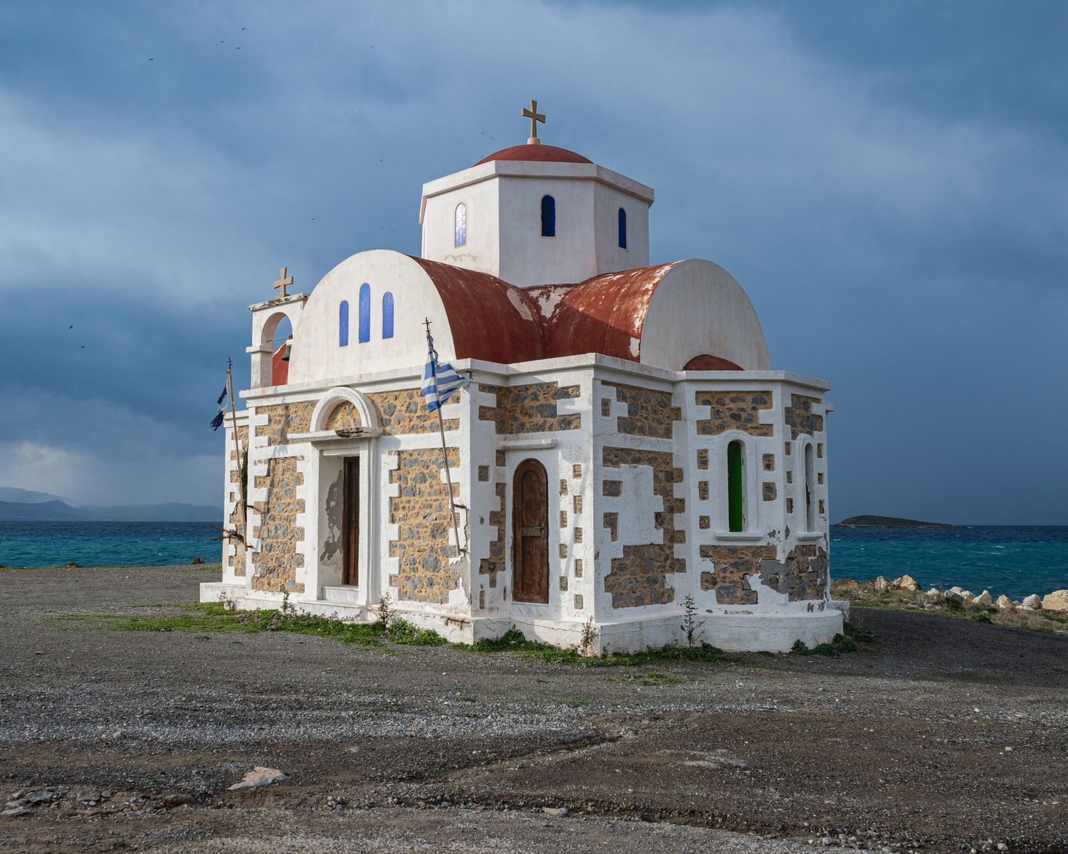 A small white Greek Orthodox church with a red dome sits beside the turquoise sea under a dramatic cloudy sky.