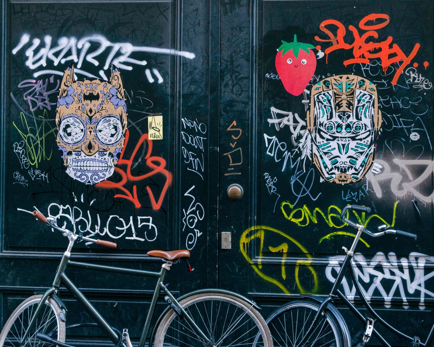 A bicycle parked against a graffiti-covered door featuring colorful sugar skull and robot face street art.