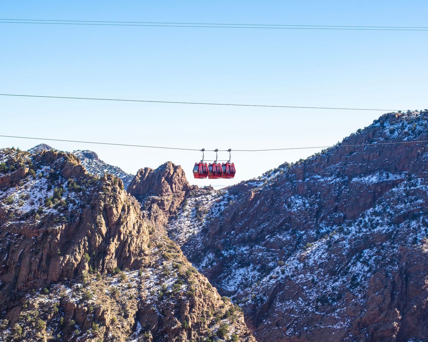 Red aerial tramway gondola glides over snow-dusted rocky canyon mountains against a clear blue winter sky.