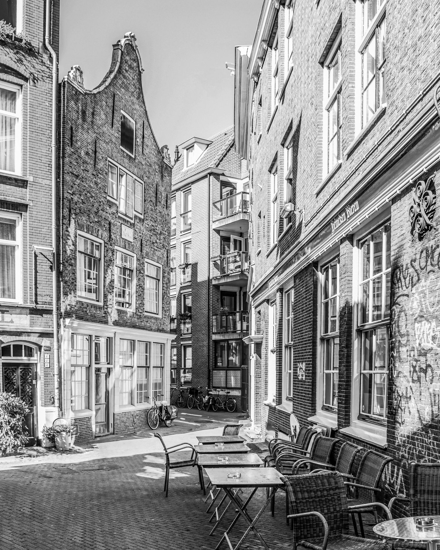Black and white photo of a quiet European alley with cafe chairs, historic brick buildings, and graffiti-covered walls.