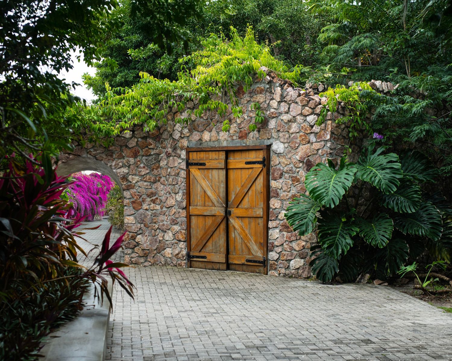 Rustic wooden double doors set in a stone wall surrounded by lush tropical greenery and colorful plants.