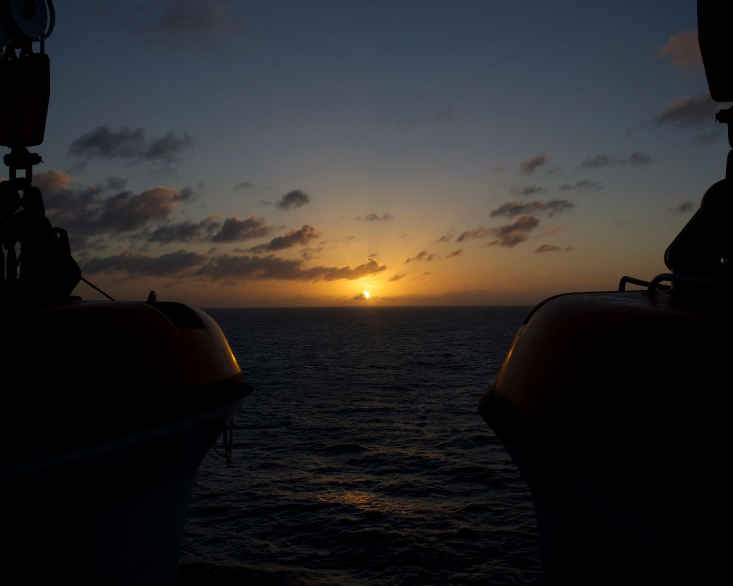 Sunset over ocean viewed between two orange lifeboats on a ship deck at dusk.
