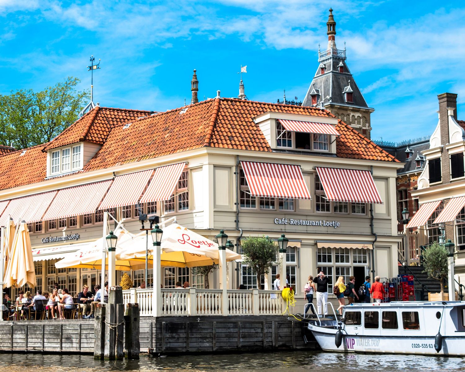 Charming Dutch canal-side restaurant with red-striped awnings, terrace seating, and a boat moored nearby on a sunny day.