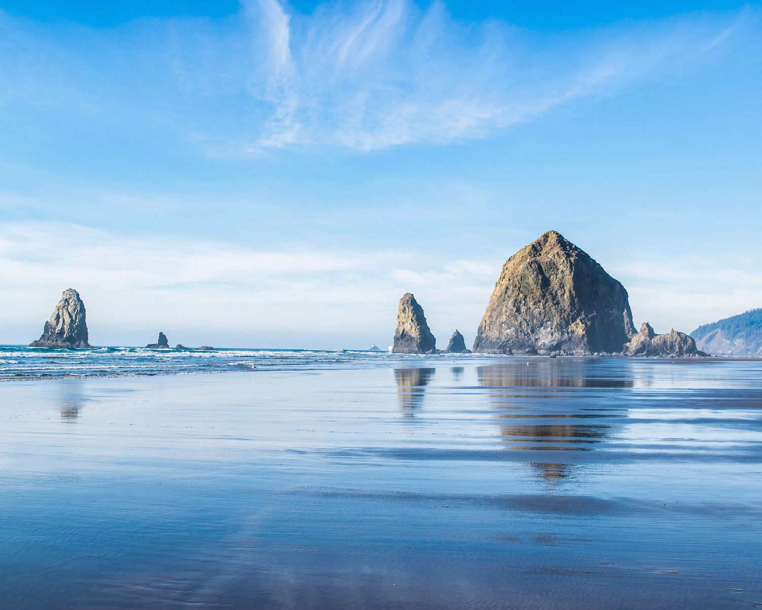 Haystack Rock and sea stacks reflected on wet sand at Cannon Beach, Oregon under a blue sky.