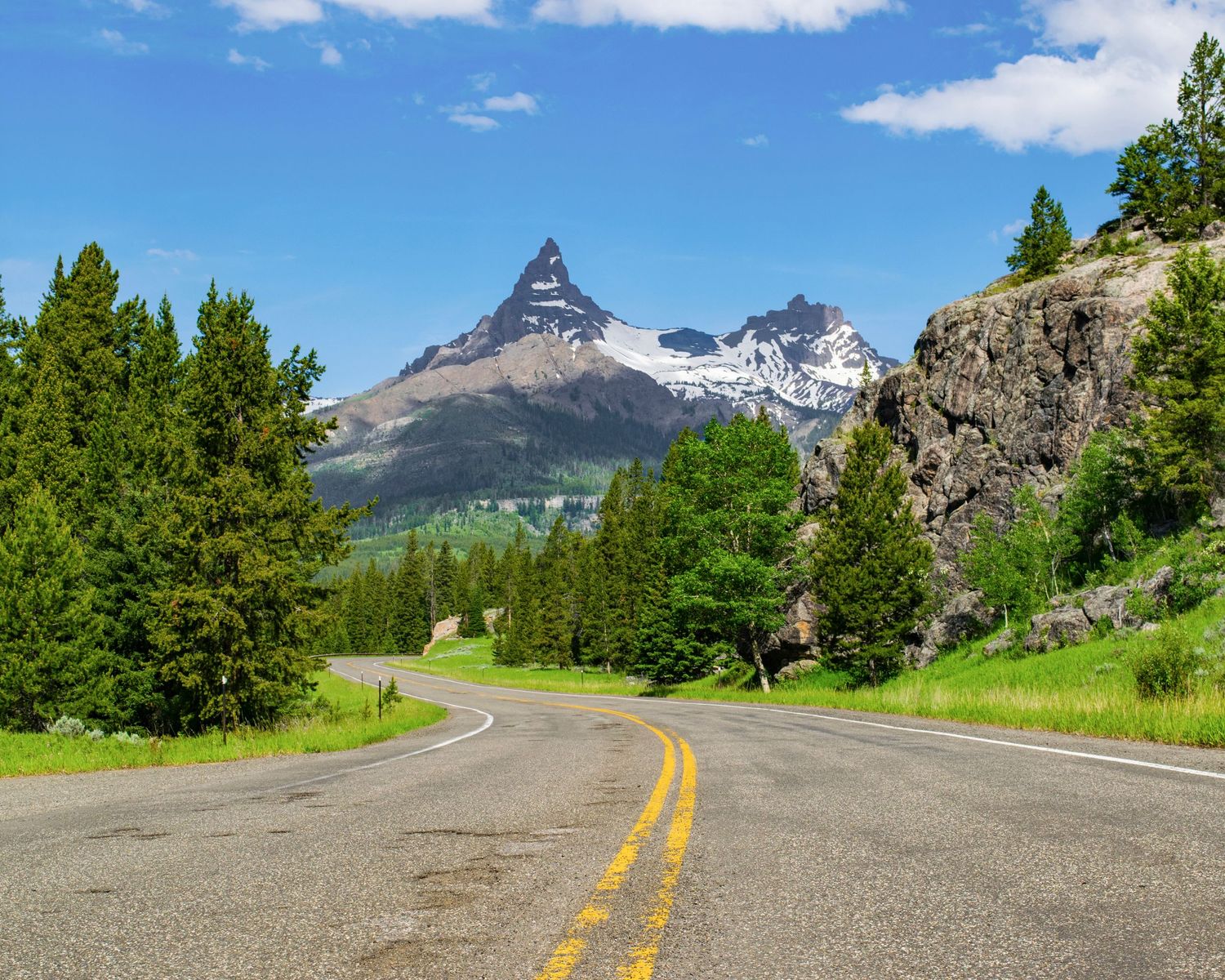 Scenic two-lane highway winding toward a sharp mountain peak with snow, flanked by evergreen trees under blue sky.