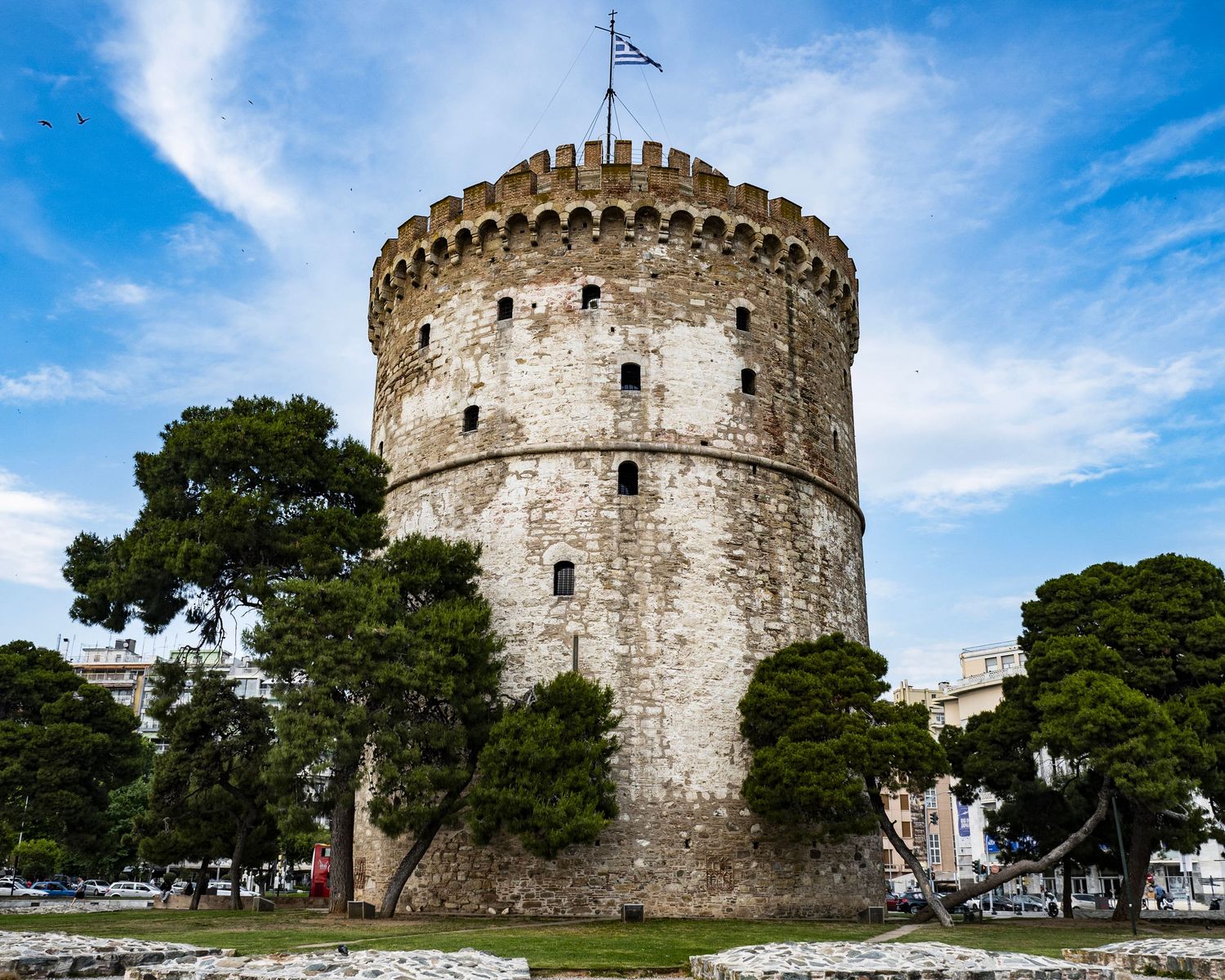 The historic White Tower of Thessaloniki, a medieval stone fortress with a Greek flag, surrounded by green trees under a blue sky.