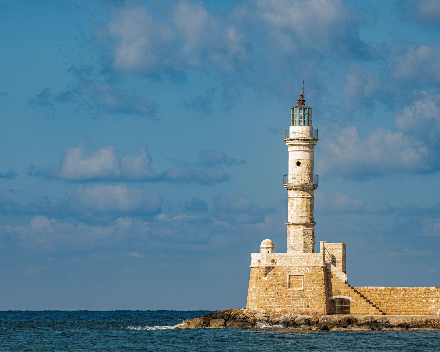 Historic stone lighthouse stands at the edge of a rocky pier in Chania, Crete, under a blue sky with clouds.