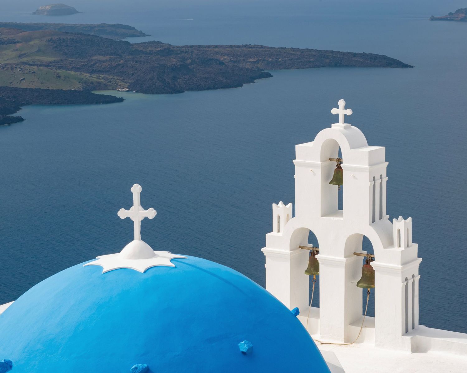 Blue-domed church with white bell tower and crosses overlooking the caldera sea in Santorini, Greece.