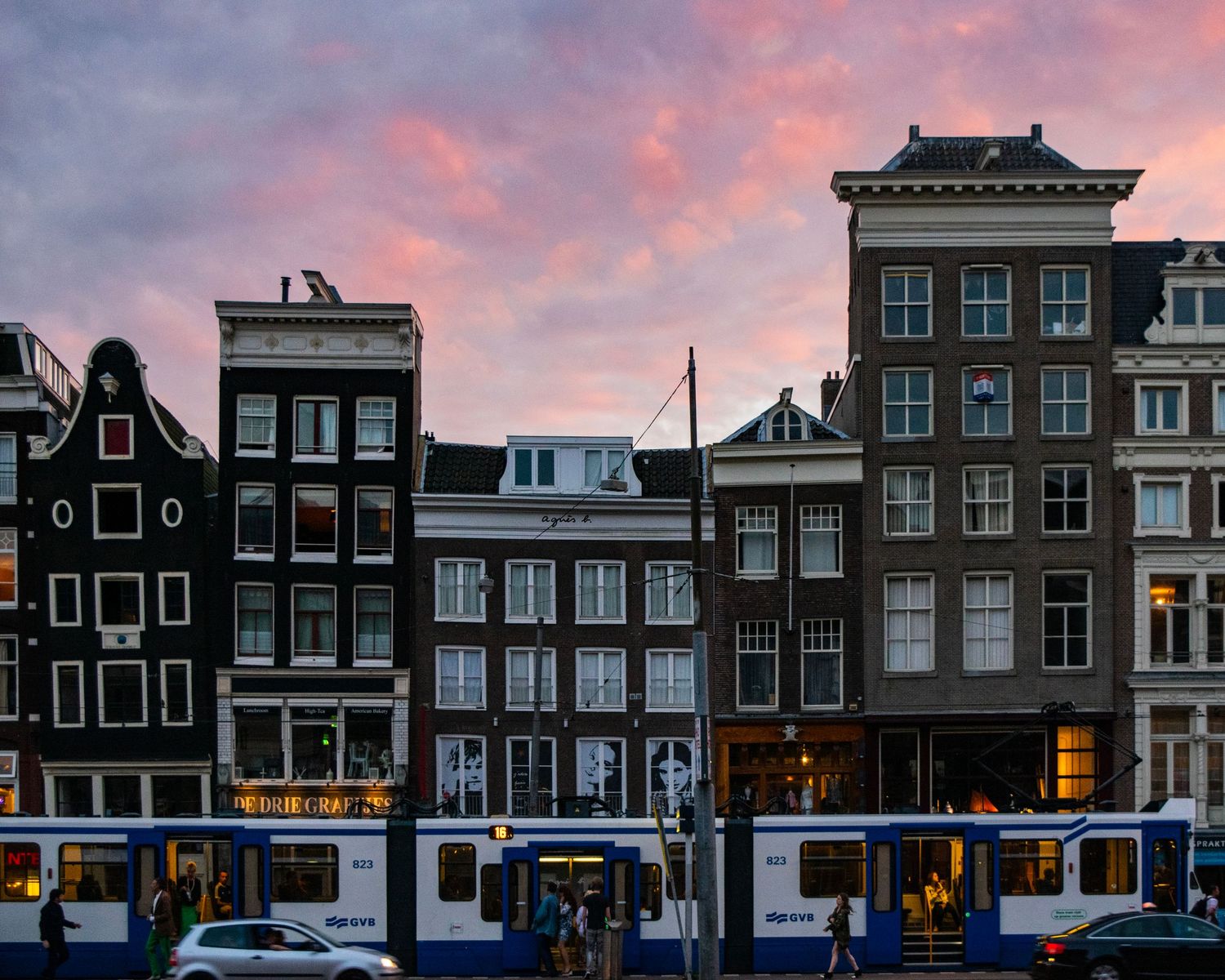 Amsterdam canal houses at dusk with a blue tram passing by under a vibrant pink and purple sunset sky.