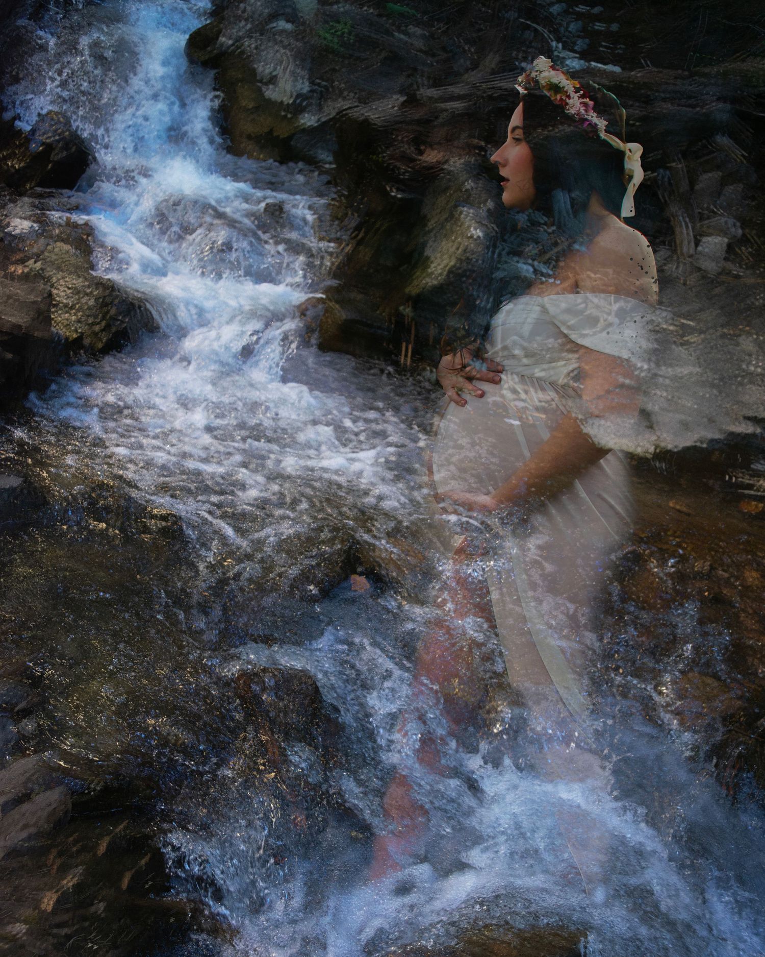 A pregnant woman in a white dress and floral crown sits ethereally among rushing waterfall rocks.