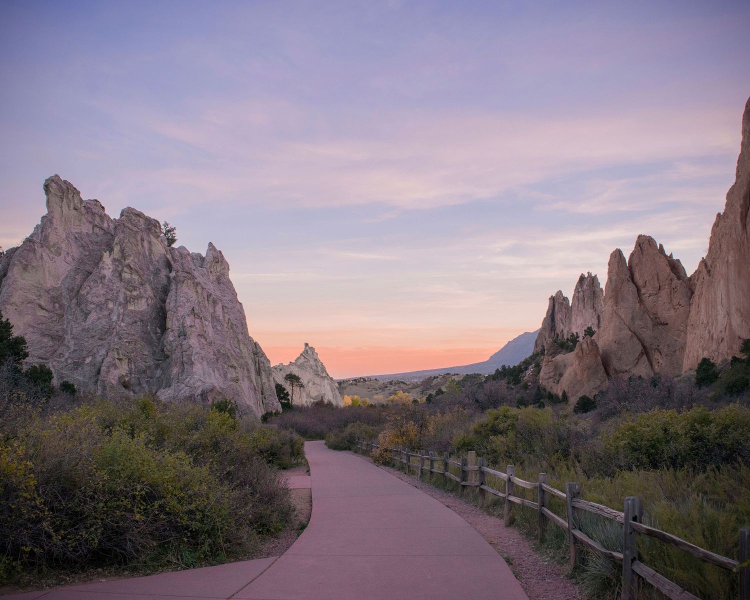 A winding paved path leads through towering red rock formations at Garden of the Gods, Colorado, under a pastel sunset sky.