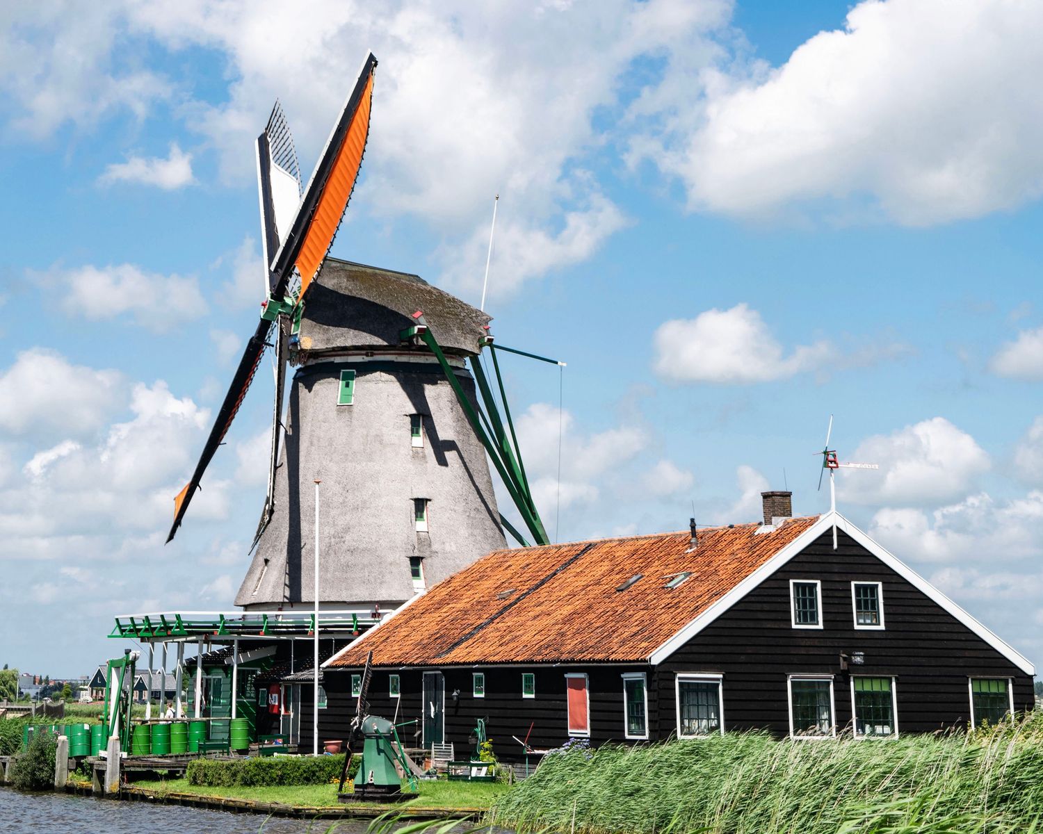 Traditional Dutch windmill beside a black wooden barn with red roof near a canal in Zaanse Schans, Netherlands.