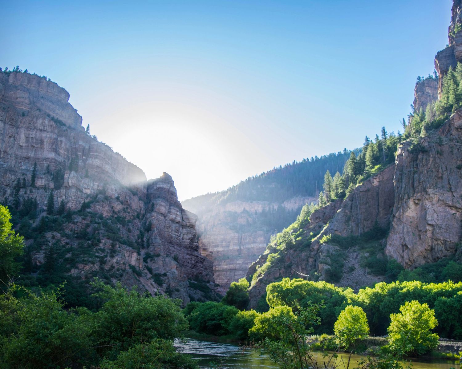 Sunlit rocky canyon with lush green trees and a winding river below under a clear blue sky.