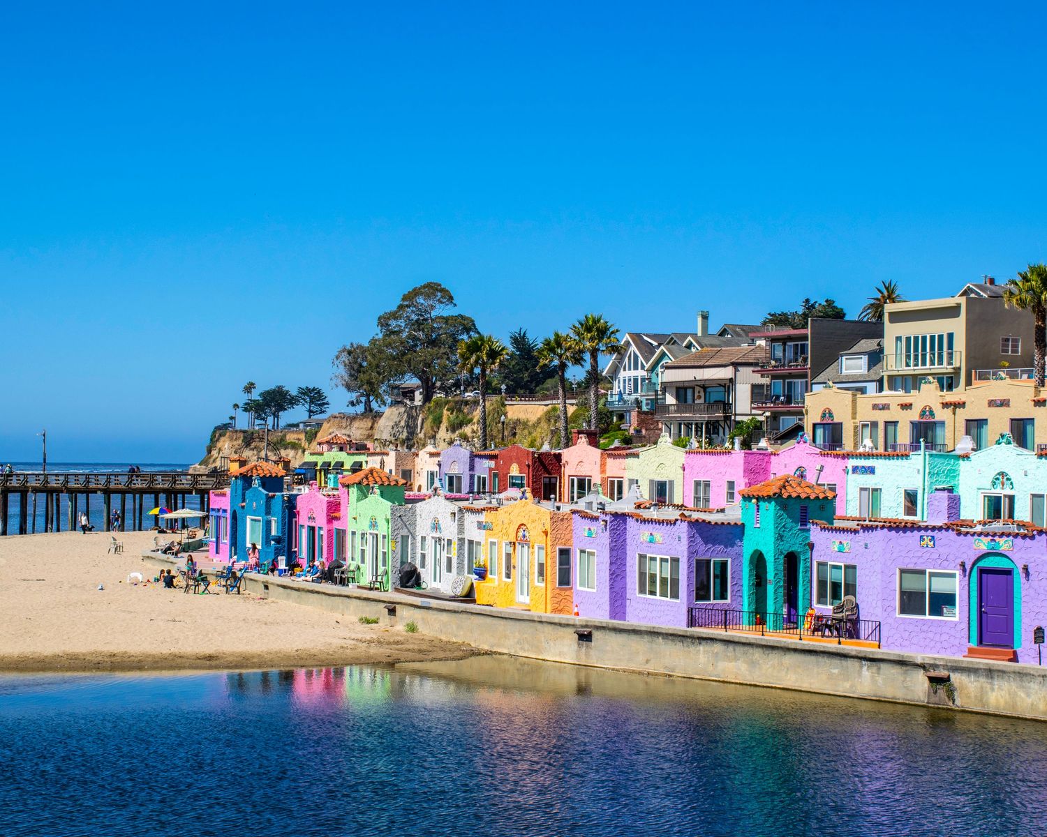 Colorful pastel beachfront buildings line the shore of Capitola Village, California, with a pier and blue sky backdrop.