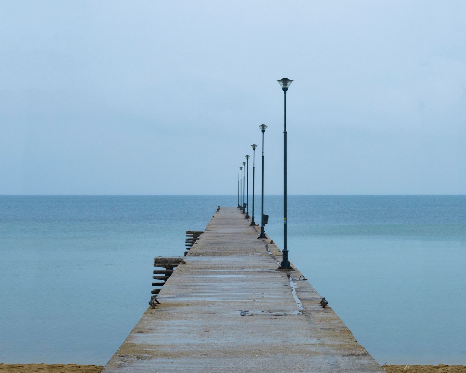 A long concrete pier with black lamp posts extends into a calm blue sea under an overcast sky.