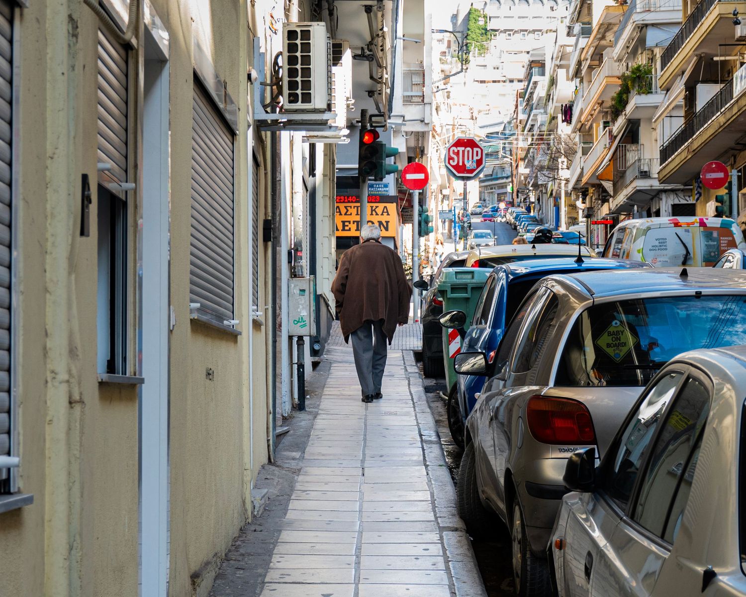 A person walks down a narrow European city street lined with parked cars and a stop sign on a sunny day.