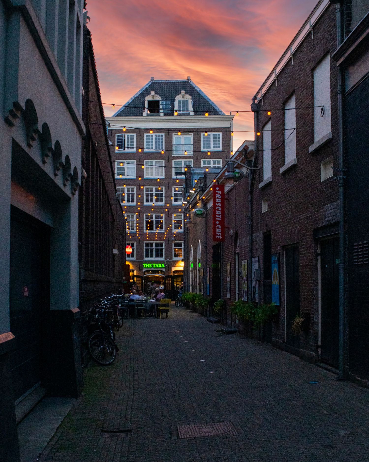 Narrow cobblestone alley in Amsterdam at sunset with dramatic red sky, brick buildings, and a green restaurant sign.