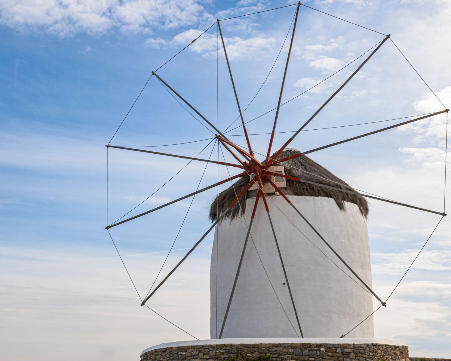 White traditional Greek windmill with wooden sail frame against a partly cloudy blue sky in Mykonos, Greece.