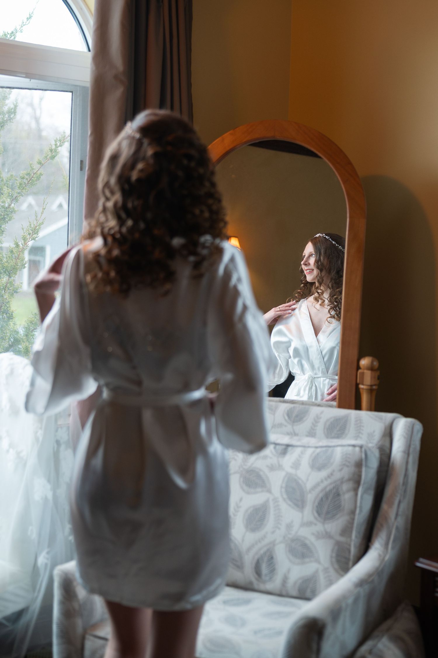 Bride in white satin robe smiling at her reflection in an arched mirror while getting ready on her wedding day.