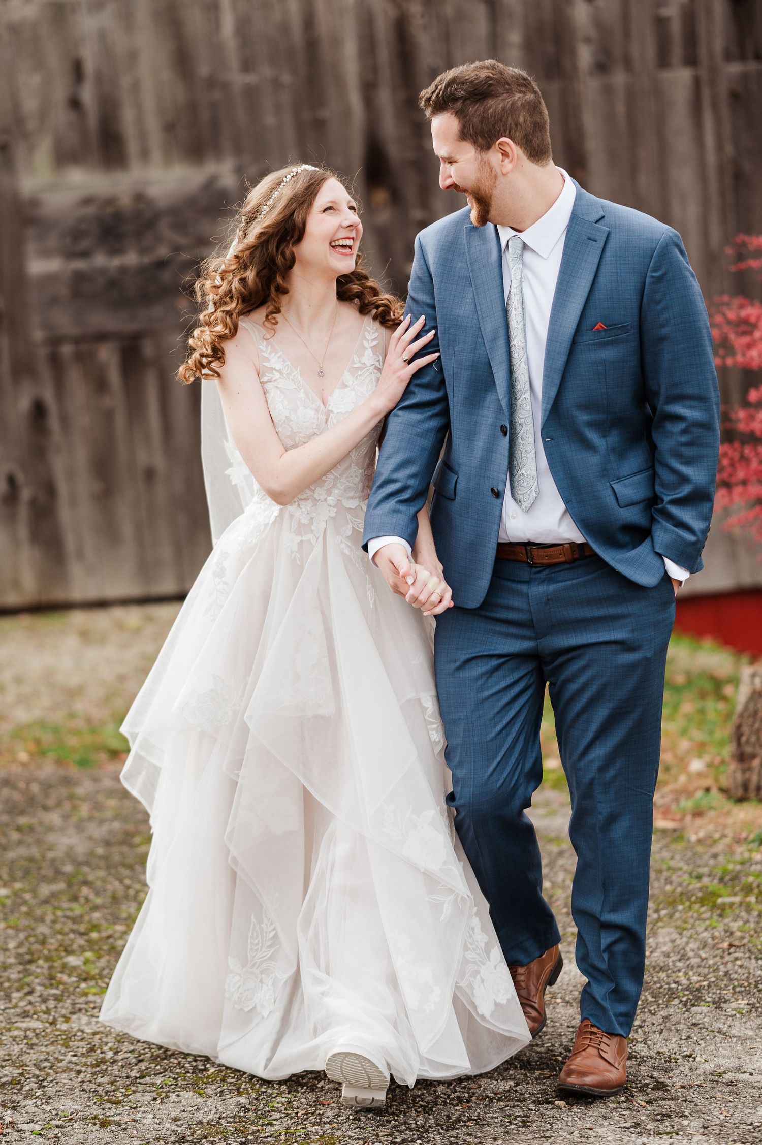 A laughing bride in a white lace gown holds hands with a groom in a blue suit during an outdoor wedding.
