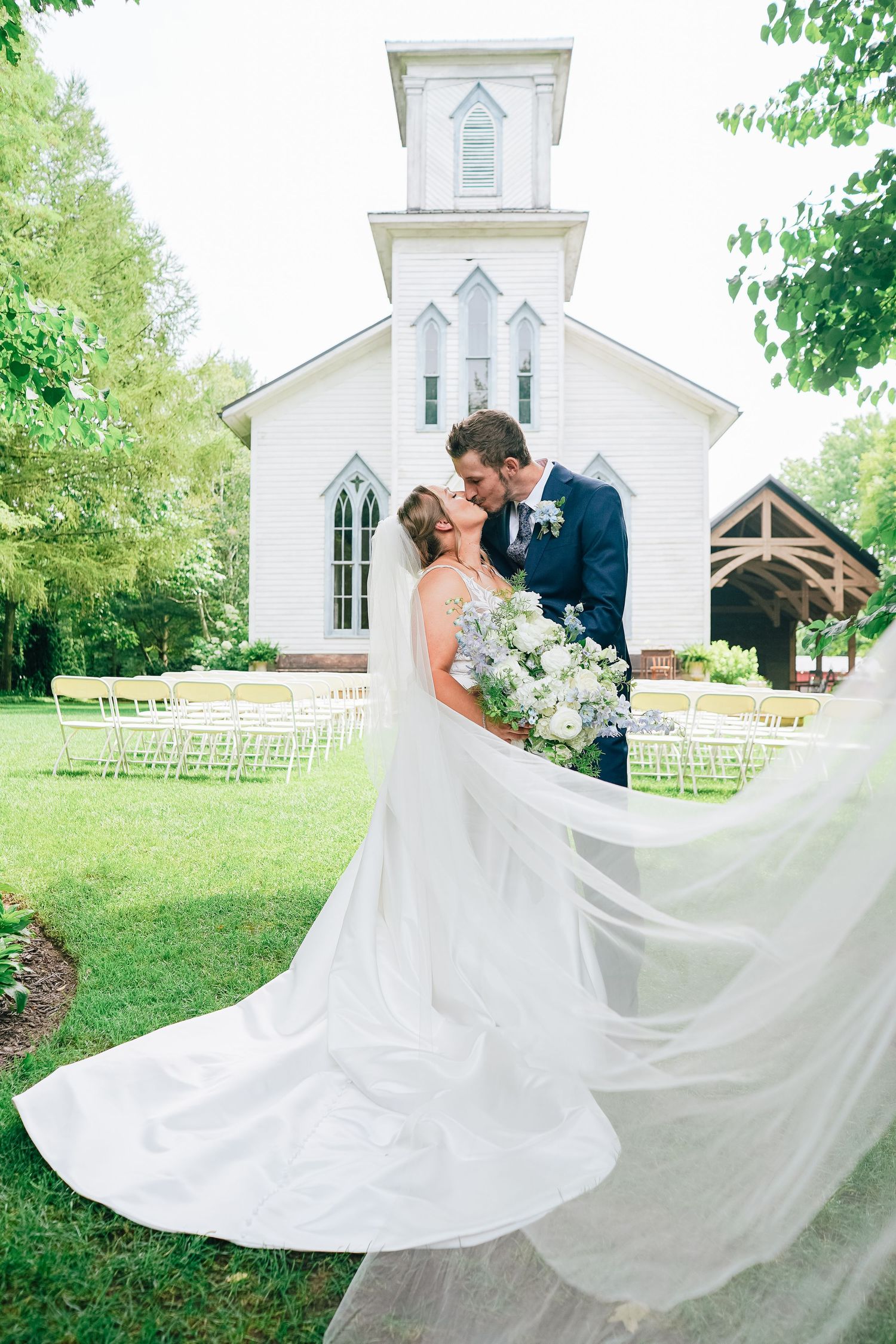 Bride and groom kissing in front of a white chapel with flowing wedding veil, surrounded by lush greenery and outdoor ceremony chairs.