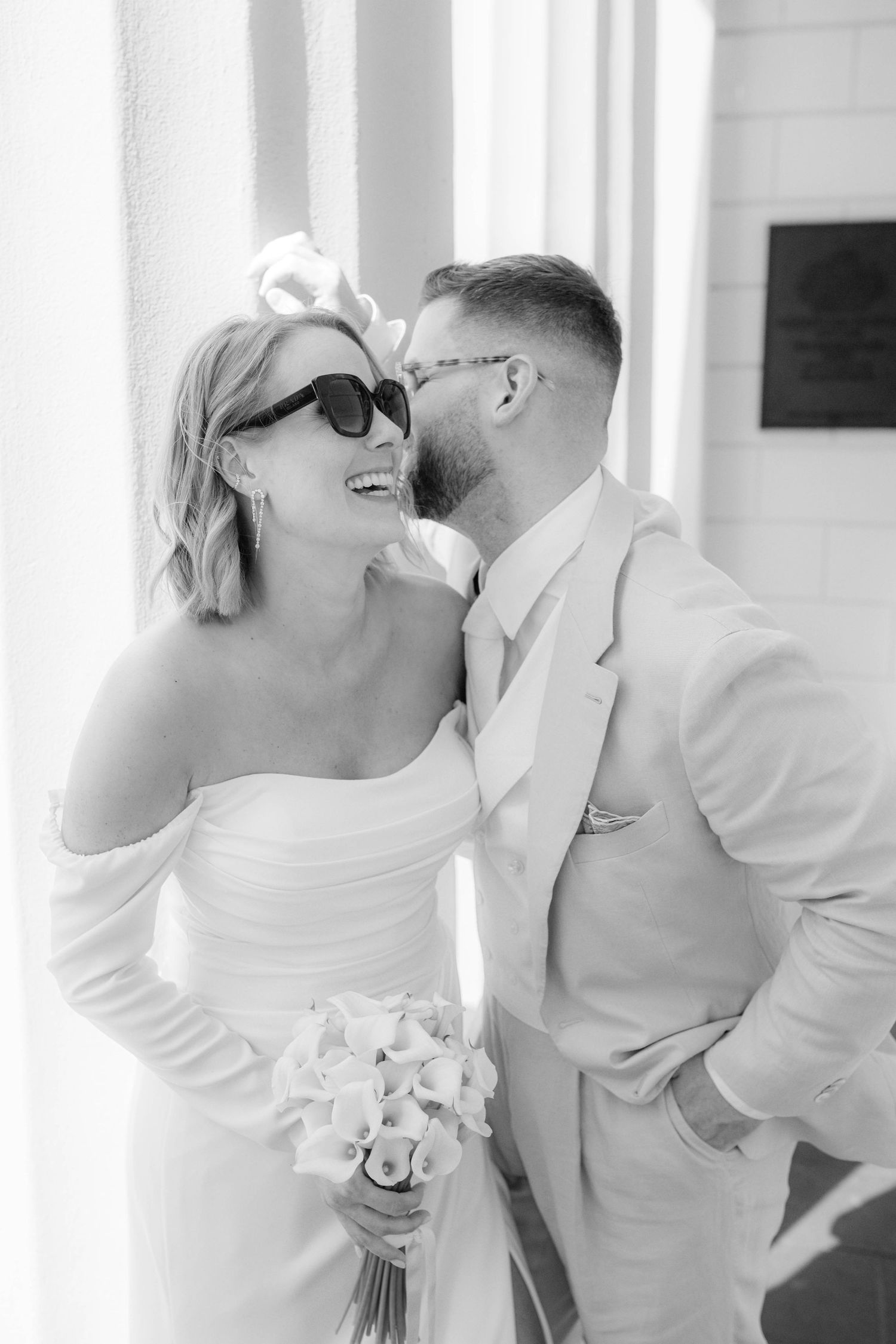 Black and white photo of a smiling bride in sunglasses holding white flowers as groom whispers in her ear.