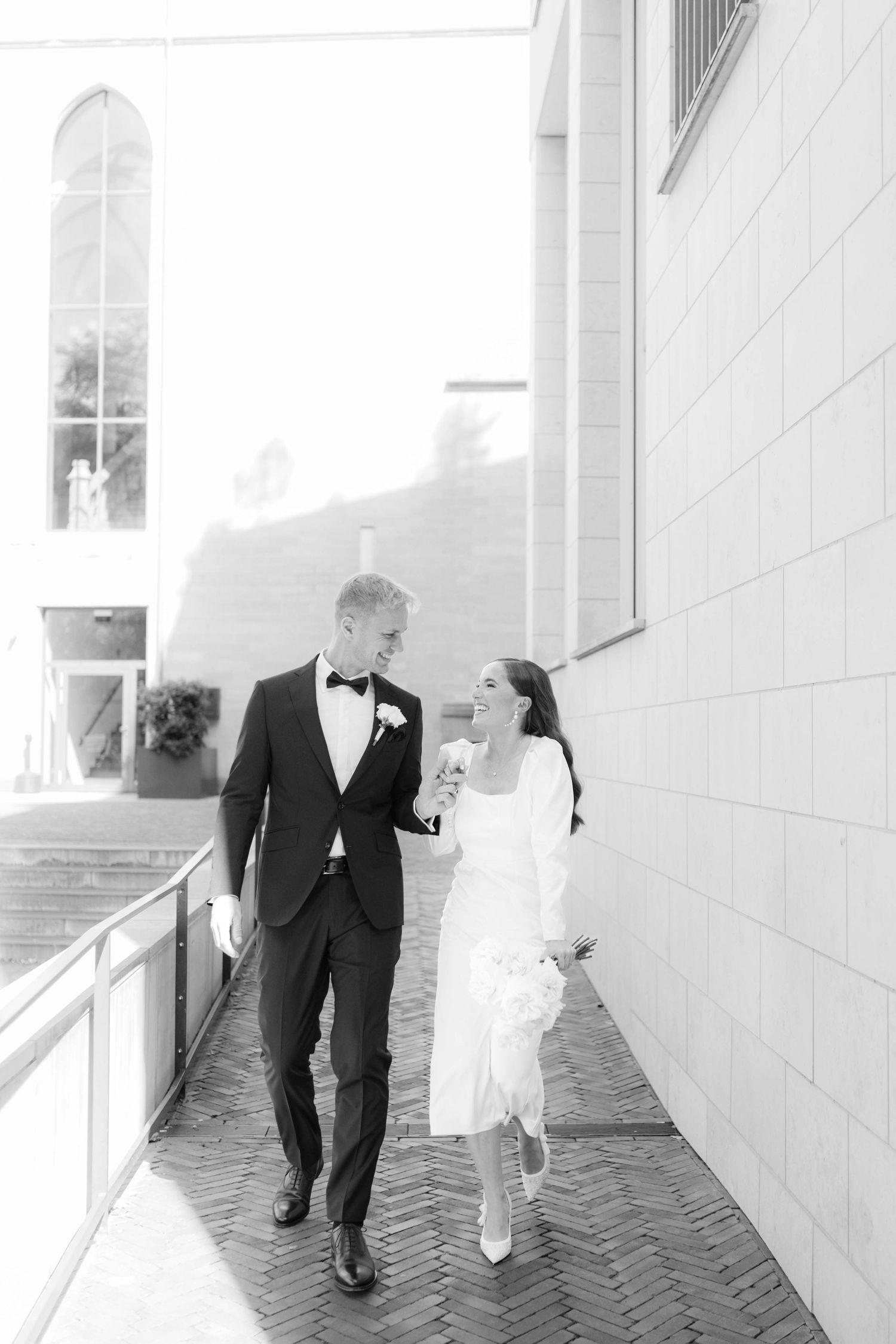 A bride and groom share a playful moment walking along a brick pathway outside a white building.