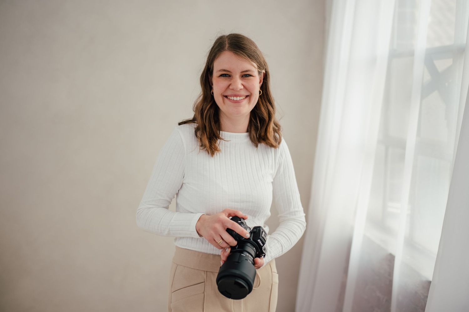 A person in white stands beside bright windows, holding a camera while taking photos in an airy indoor space.