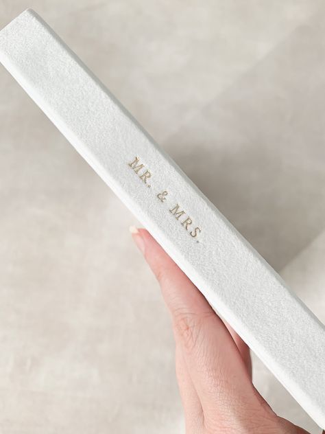 Hand holding white wedding album with gold embossed text against neutral background.