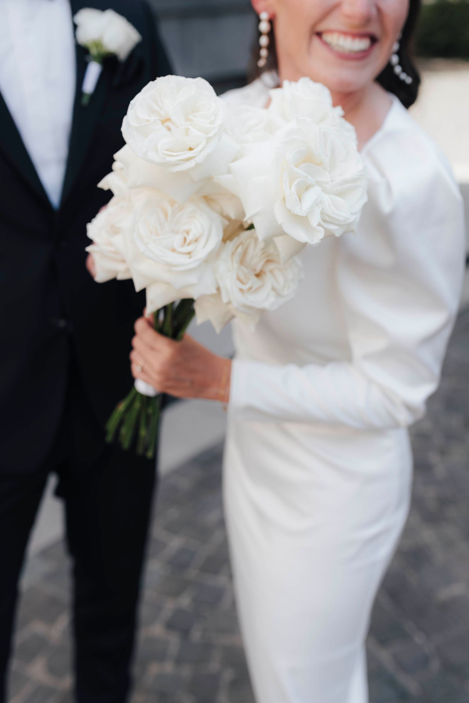 Bride in white gown holding elegant white rose bouquet beside groom in black tuxedo at wedding.