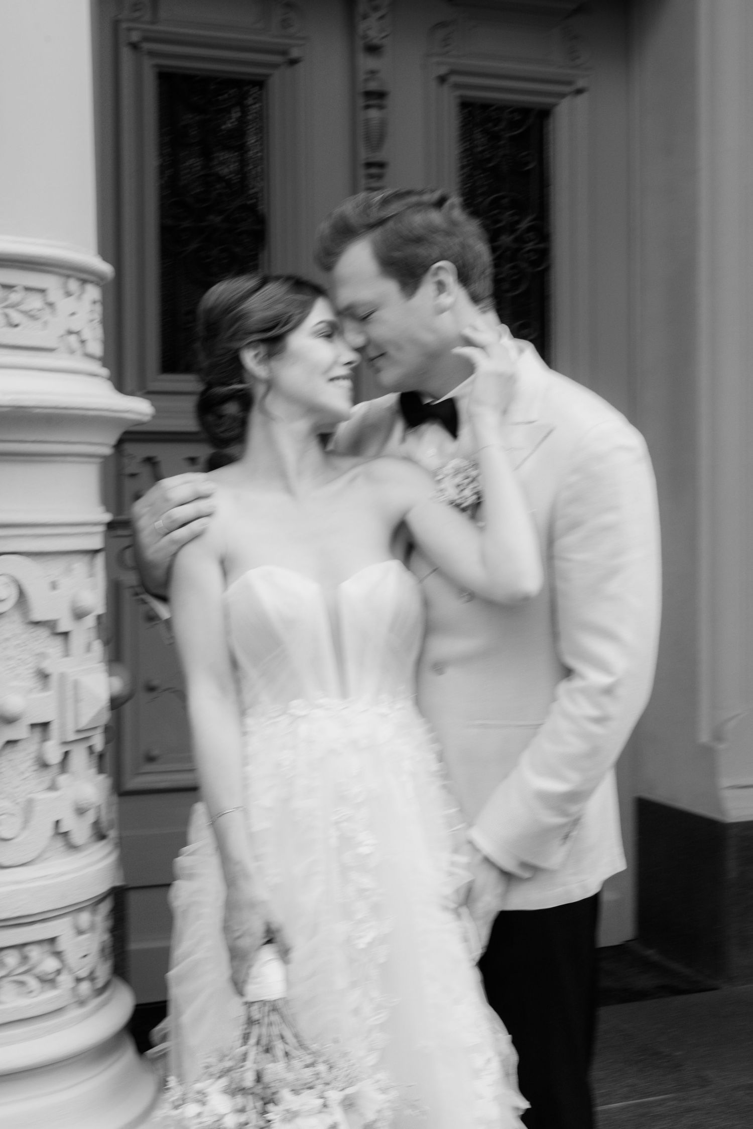 Black and white photo of a bride and groom sharing a romantic moment by an ornate column at their wedding.