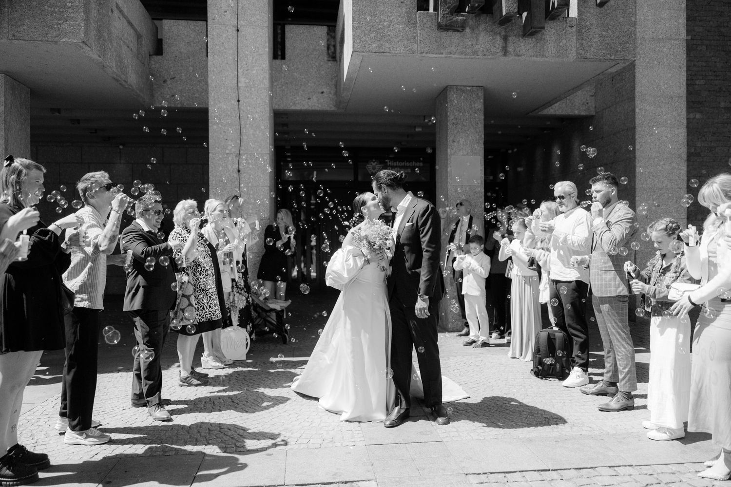 A black and white photo of a wedding couple kissing outside a building while guests blow bubbles around them.