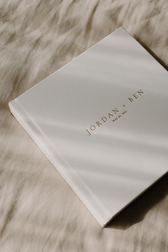 Minimalist white wedding album cover with gold text 'JORDAN + BEN' on textured fabric.