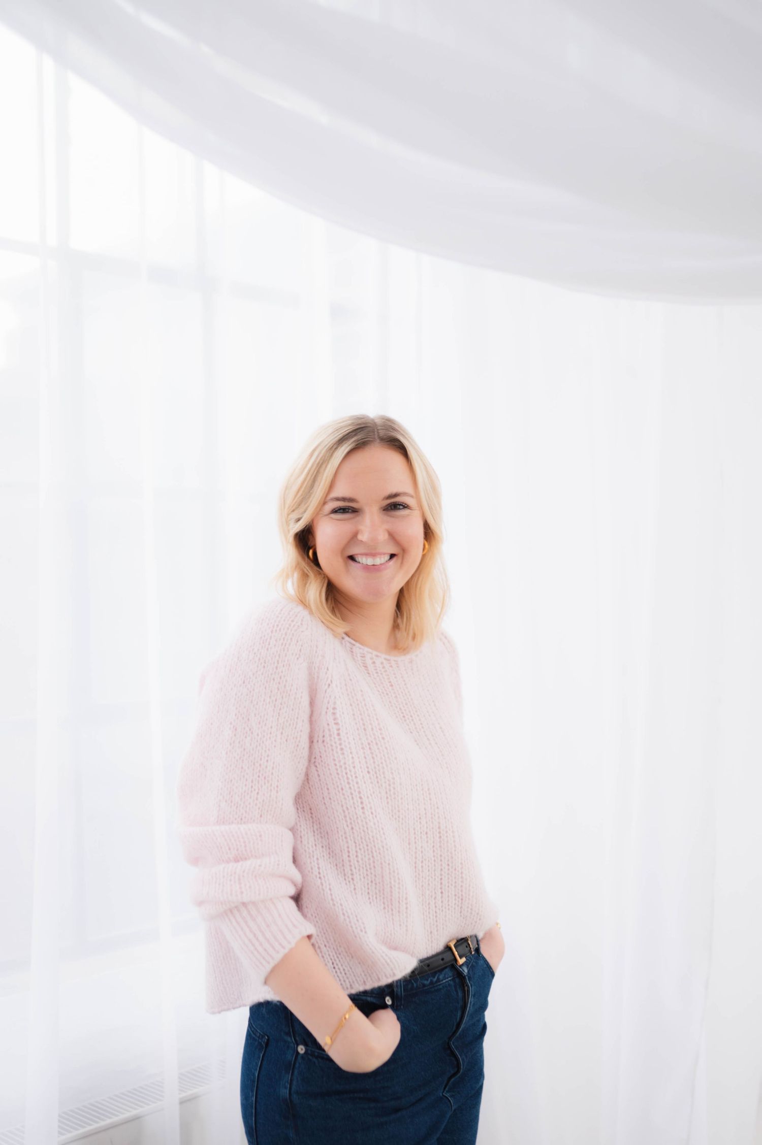 A person wearing a soft pink sweater and dark jeans poses against a bright white background in a studio setting.