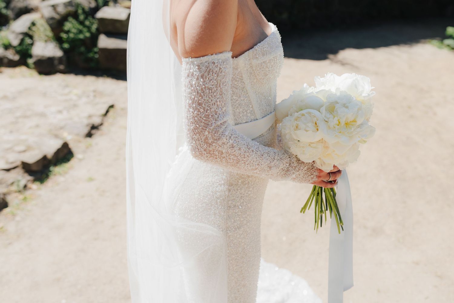A bride in a fitted lace wedding dress holds a delicate white and blue bouquet in sunlight.