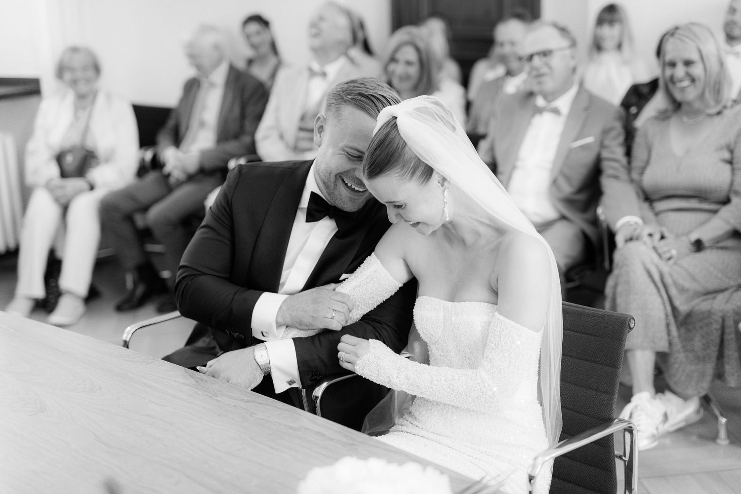 A bride and groom sit at a wedding ceremony table, signing documents while guests look on in black and white.