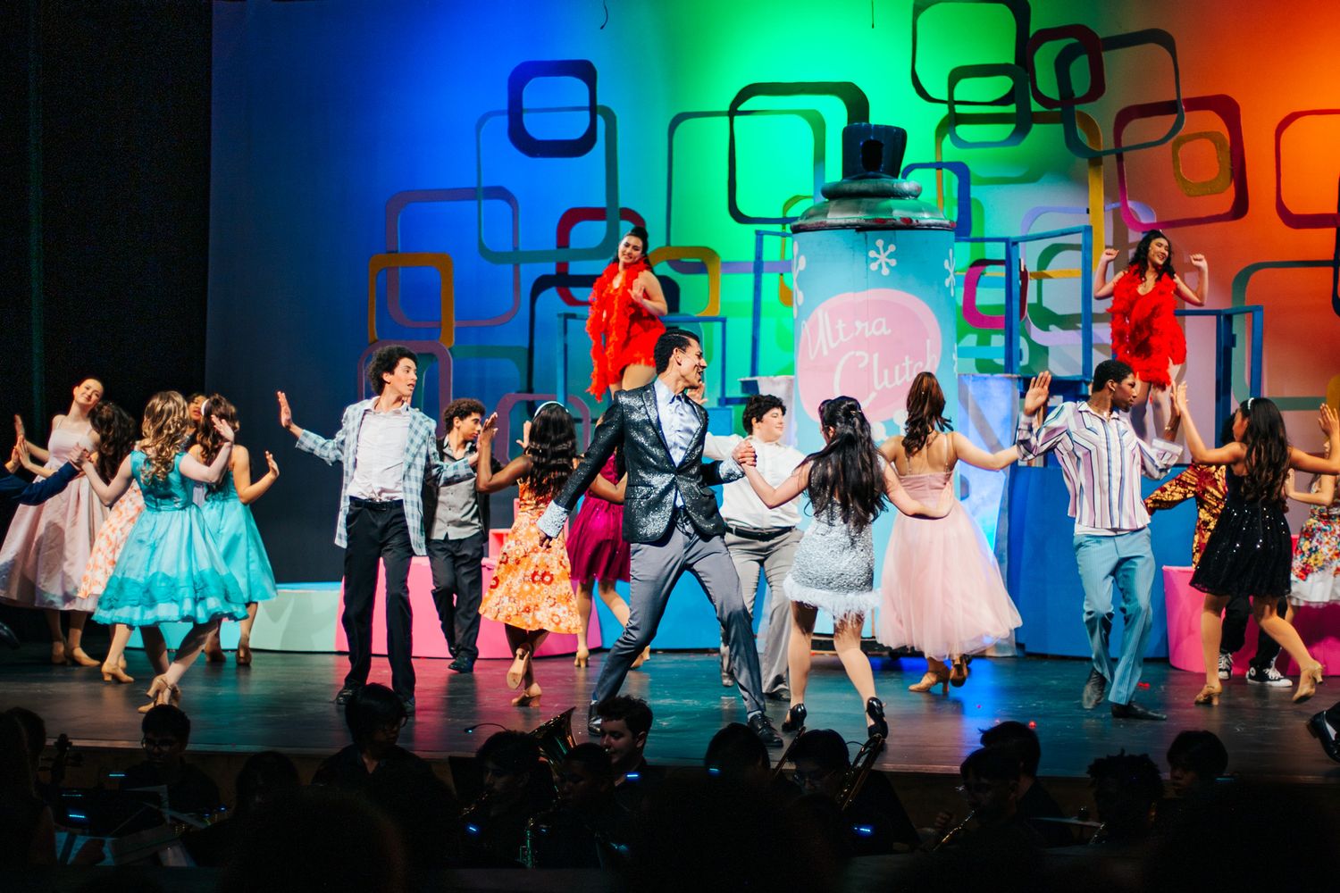 A colorful stage performance with dancers in 1950s style clothing performing against a vibrant geometric backdrop.