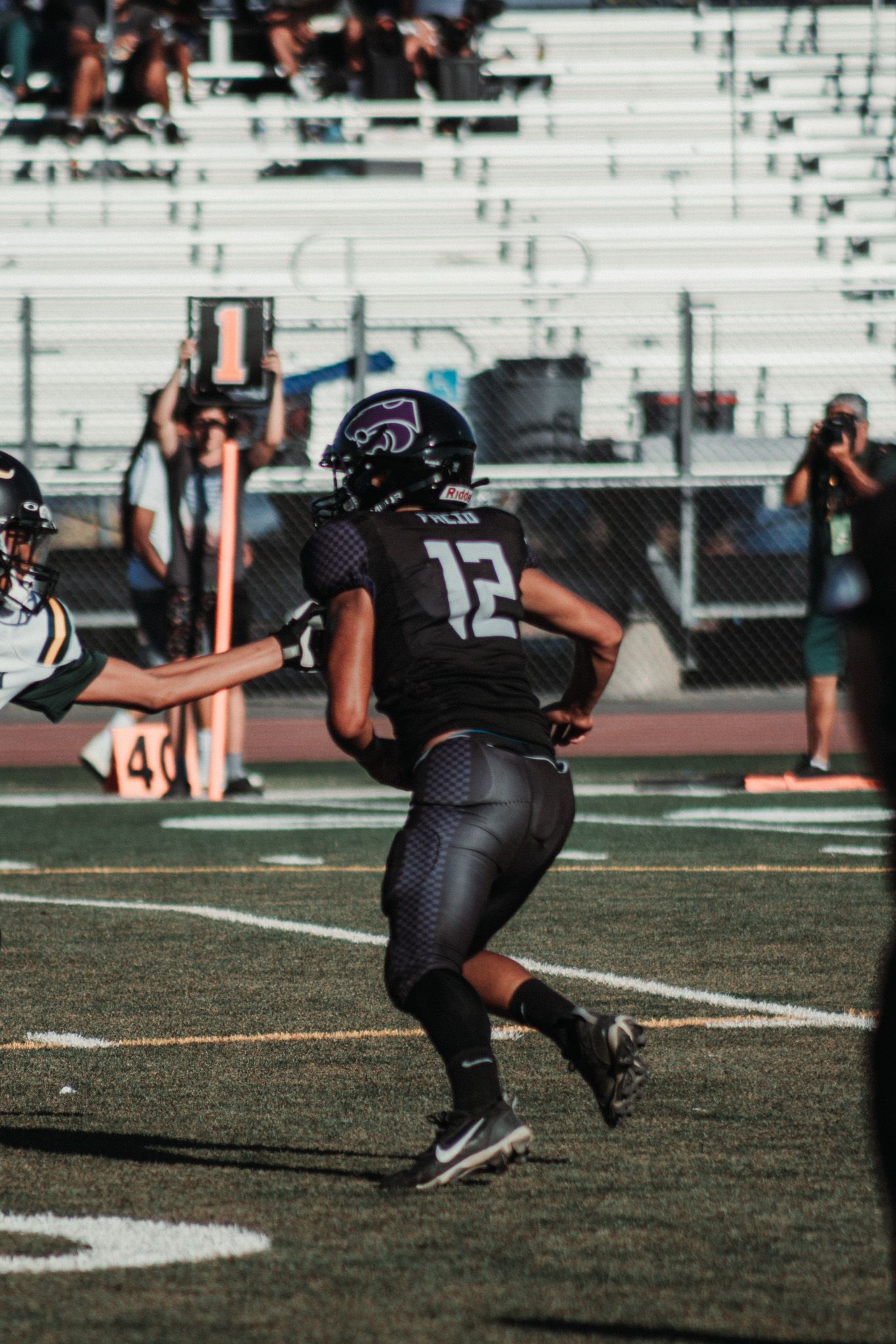 Football player in black jersey number 12 runs on field during game with stadium bleachers in background.