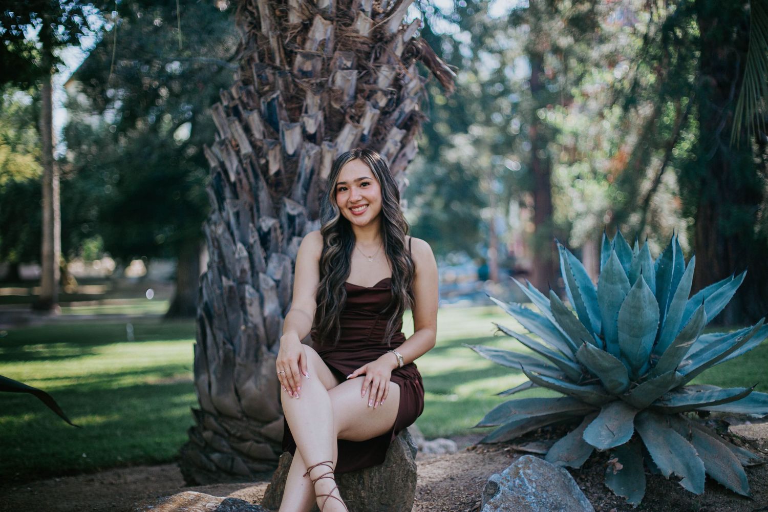A woman in a brown dress poses outdoors next to a large tree trunk and blue agave plant in a sunny garden setting.