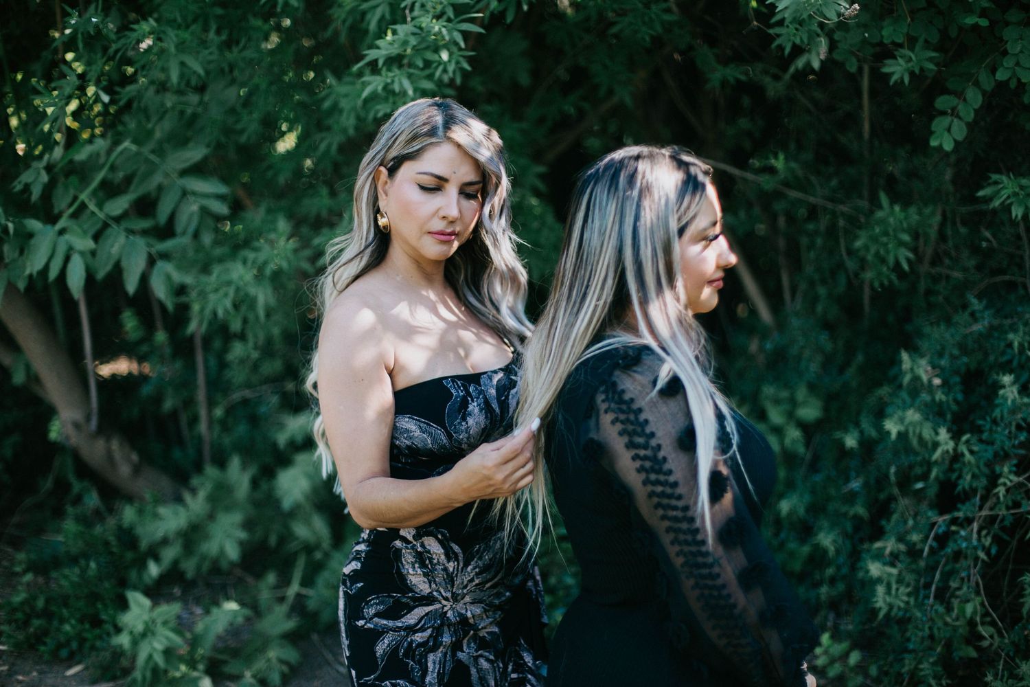 Two women in contrasting black outfits pose together against lush green foliage in an artistic outdoor portrait.