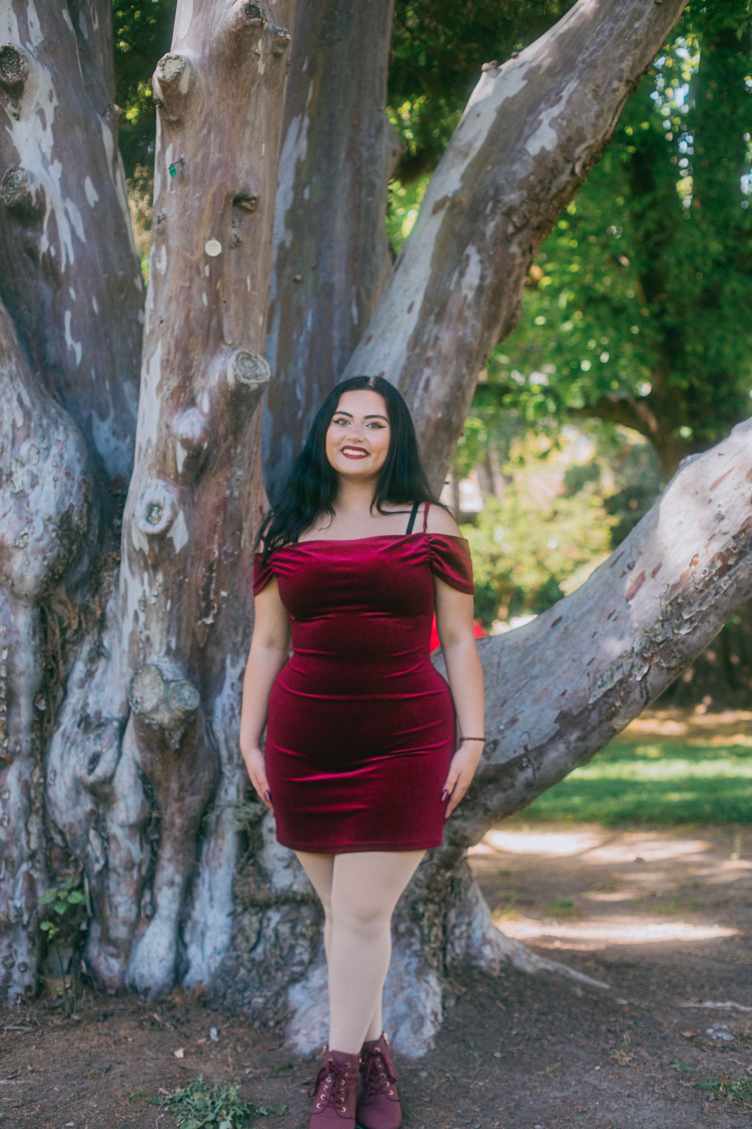 A burgundy velvet mini dress is worn against a backdrop of large tree trunks in an outdoor setting.