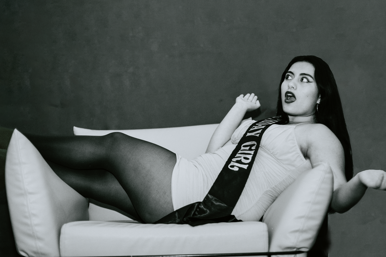 A dramatic black and white photo shows someone reclining theatrically on a white couch wearing dark stockings.