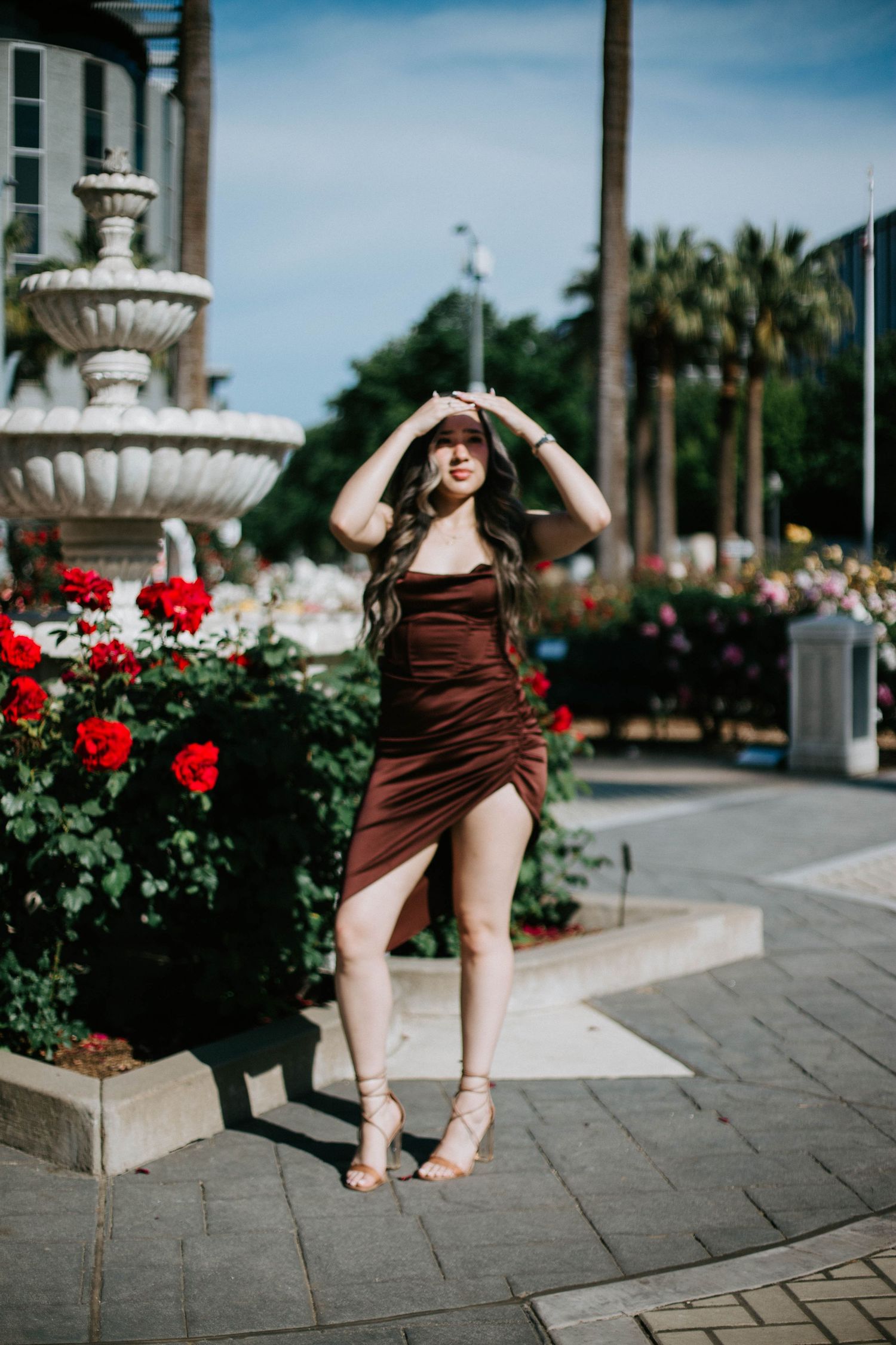 Woman in brown dress poses near ornate fountain and red flowers in outdoor urban setting with palm trees.