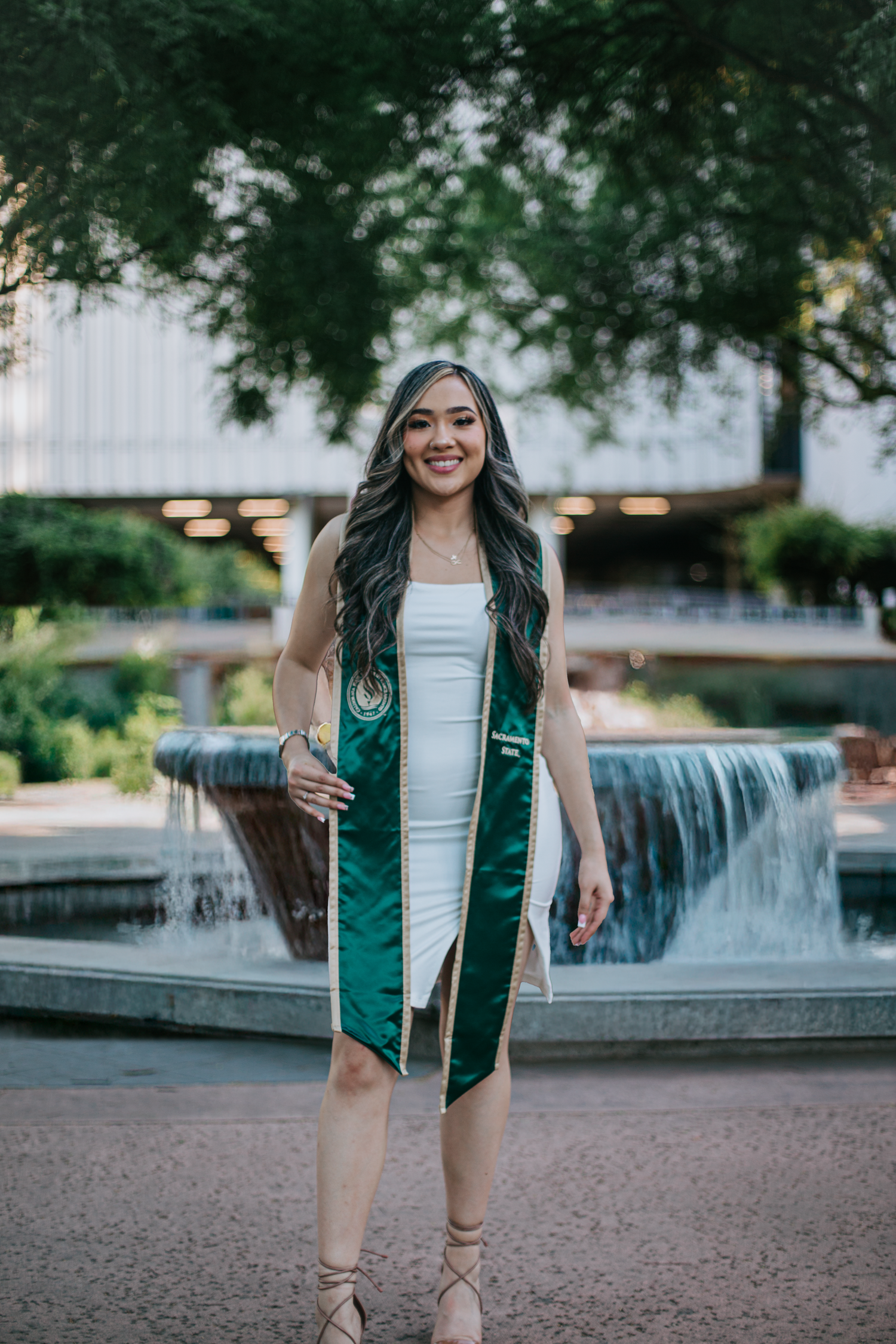Graduate in white dress and green graduation stole poses with a smile in front of an outdoor fountain.
