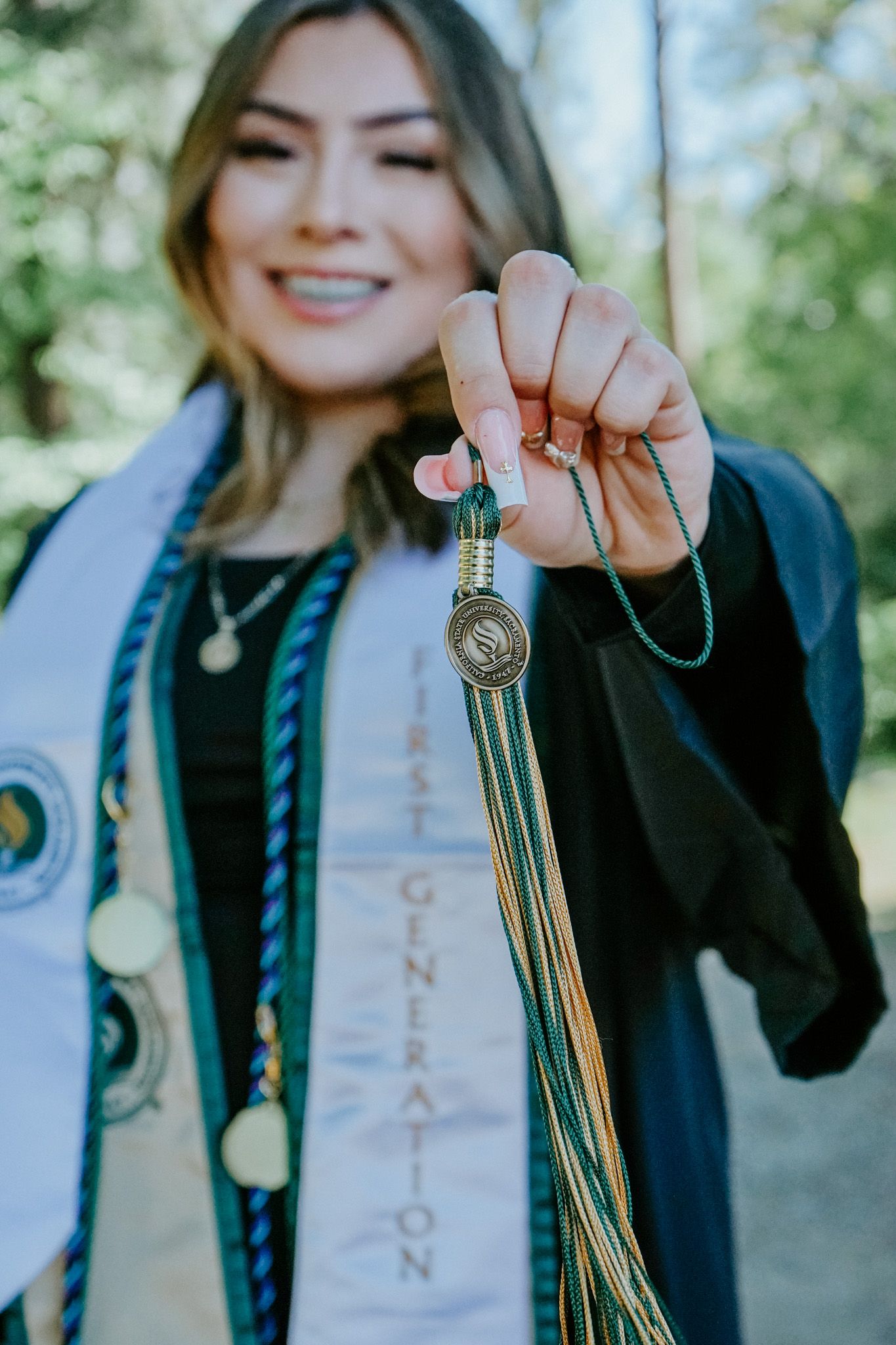 Graduate displays tassel and graduation regalia while wearing a black gown with decorative sash in outdoor setting.