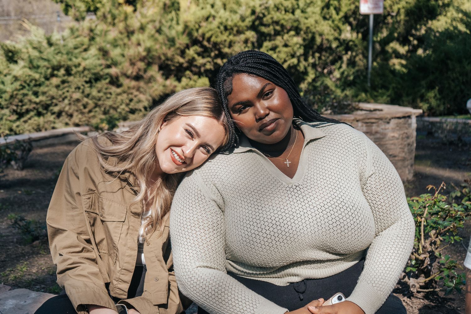Two friends sit together outdoors on a sunny day, one wearing a tan jacket and the other in a cream sweater.