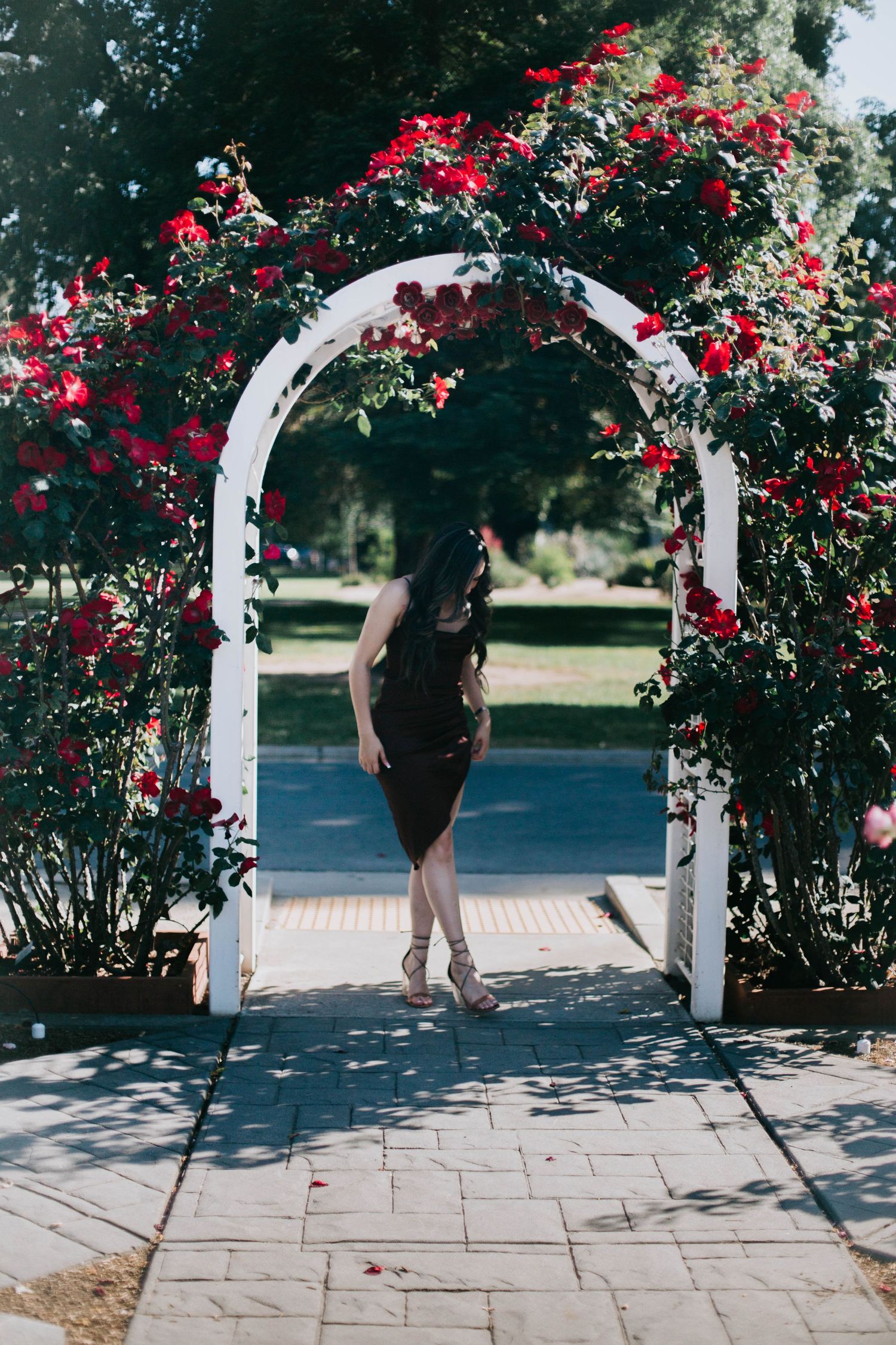 White garden arch covered in vibrant red climbing roses frames a figure on a stone pathway in a summer garden.